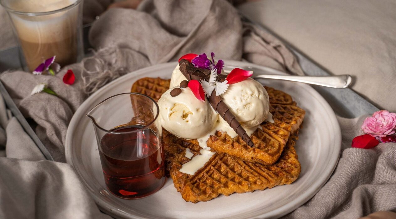 3 pieces of waffles topped with white ice-cream, chocolate and flower pettals and a jar of brown liquid on a white round plate on cloth with a cup of drink