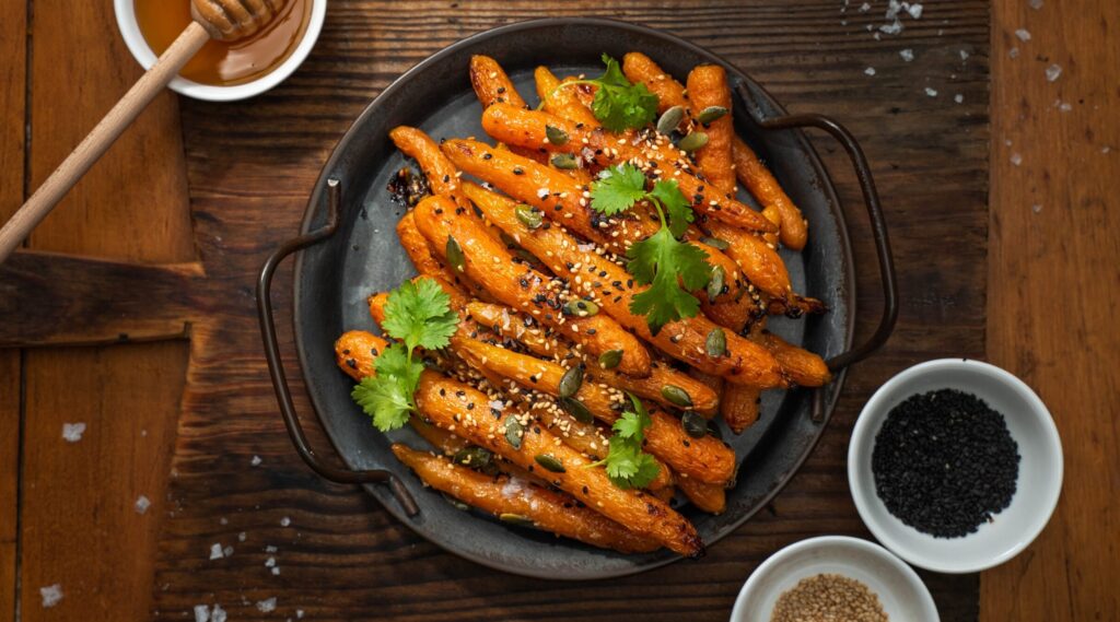 Cooked whole baby carrots on a round black skillet on wooden board, pots of honeyand white and black sesame seeds