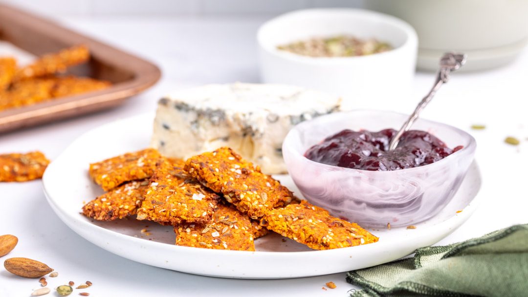 Several orange-coloured crackers on a plate with cheese and a bowl of purple jam.