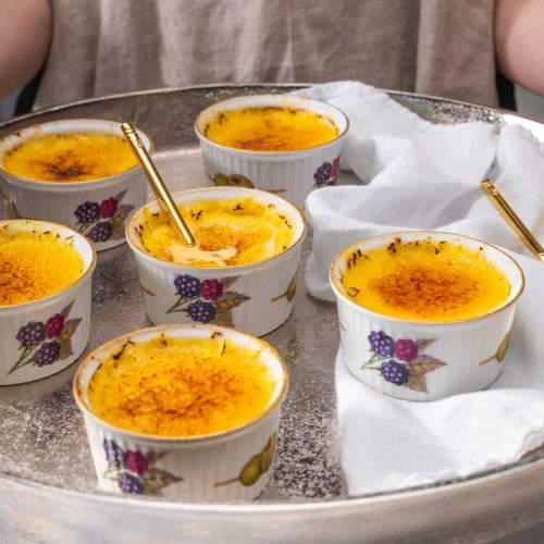 Six ramekins of creme brulee on a round silver tray.