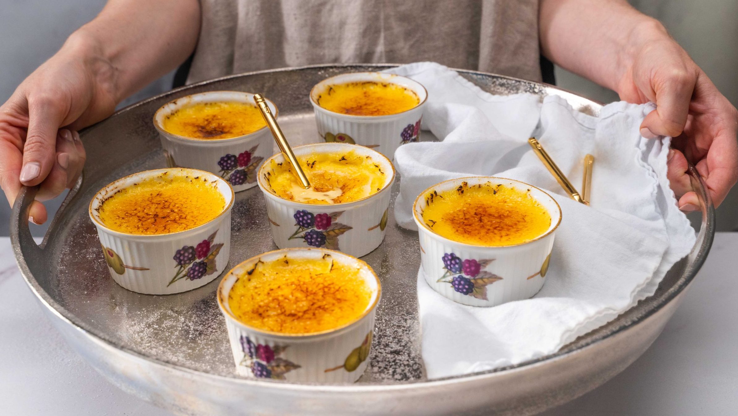 Six ramekins of creme brulee on a round silver tray.