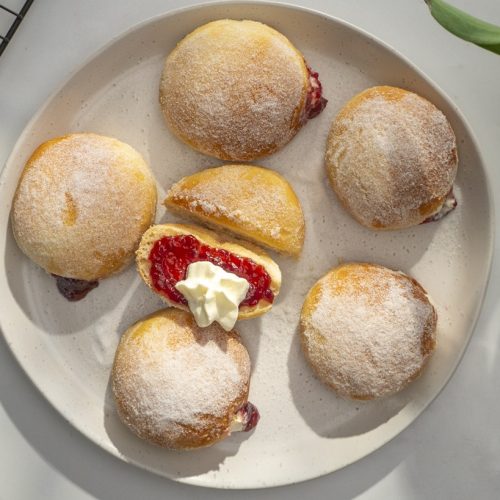 Six sugar dusted round buns on a white plate , the middle one is cut in half and showing red jam and cream inside.