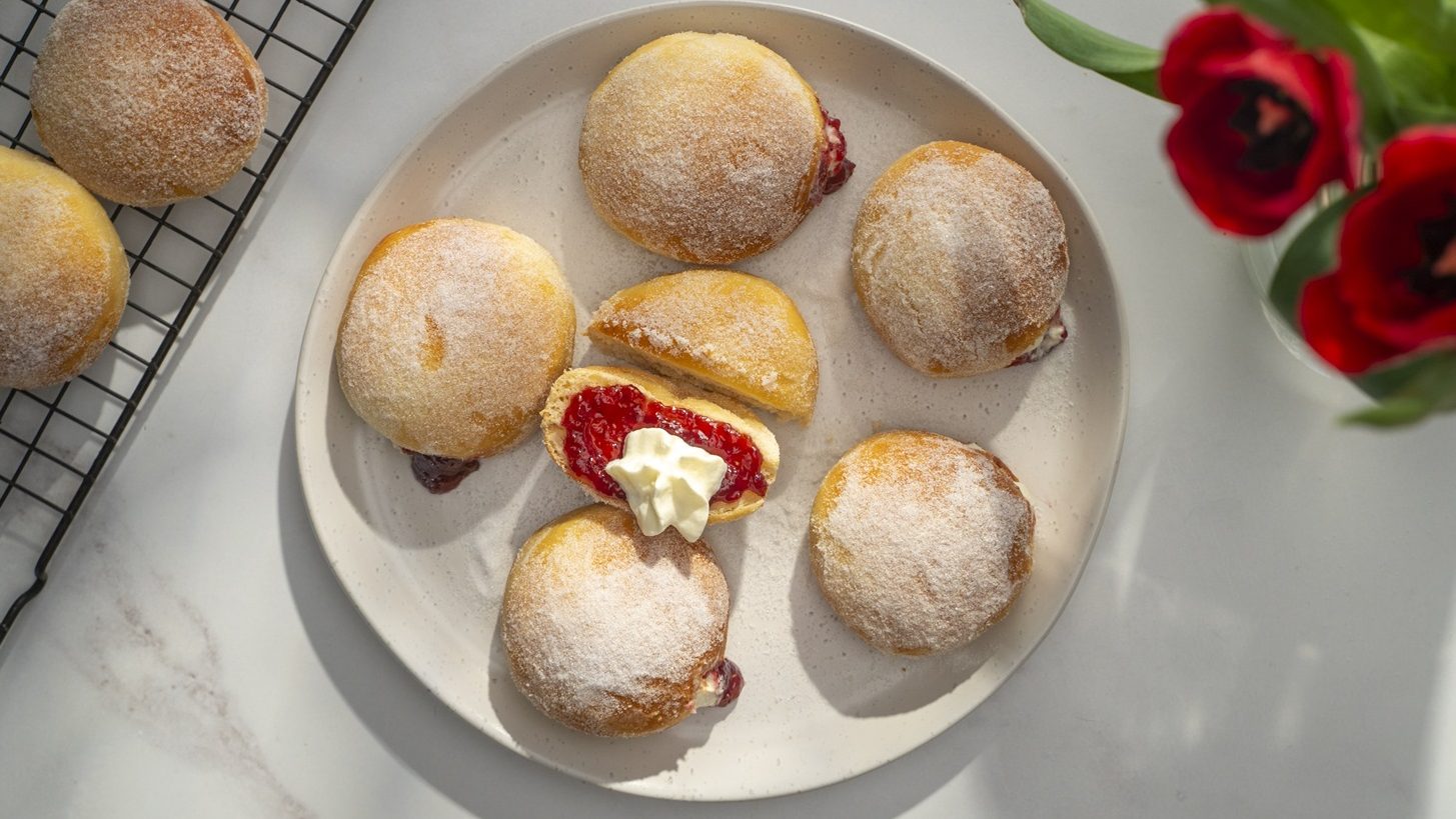 Six sugar dusted round buns on a white plate , the middle one is cut in half and showing red jam and cream inside.
