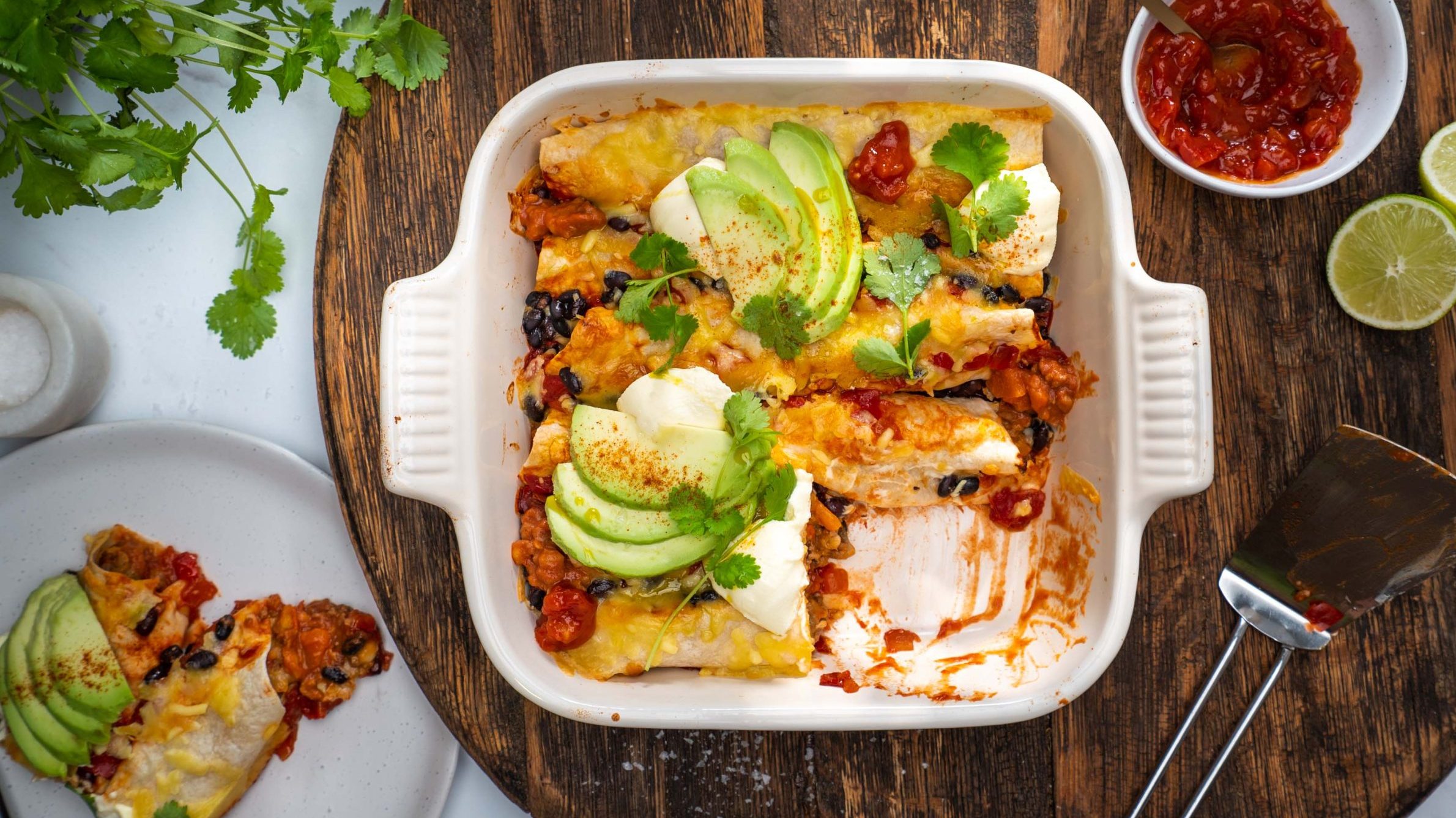 Baked enchiladas topped with sliced avocado and sour cream in square dish with a portion served on a plate.