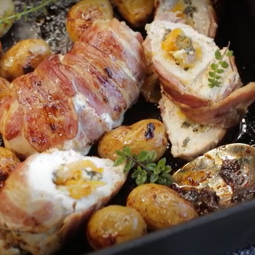 Apricot stuffed and bacon wrapped chicken breasts and baby potatoes on oven tray with a serving spoon.