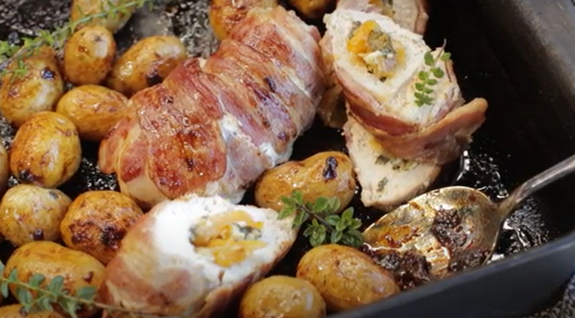 Apricot stuffed and bacon wrapped chicken breasts and baby potatoes on oven tray with a serving spoon.
