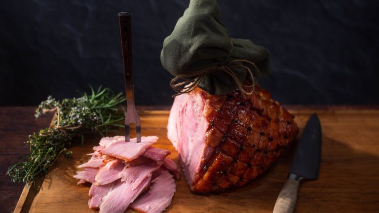 Baked whole leg ham with some sliced pieces on a wooden board.