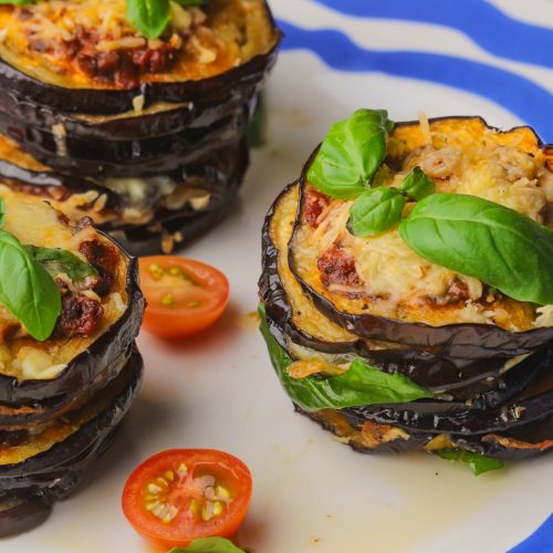 Three piles of aubergine slice stacks with basil leaves on top on a plate with cherry tomato halves.