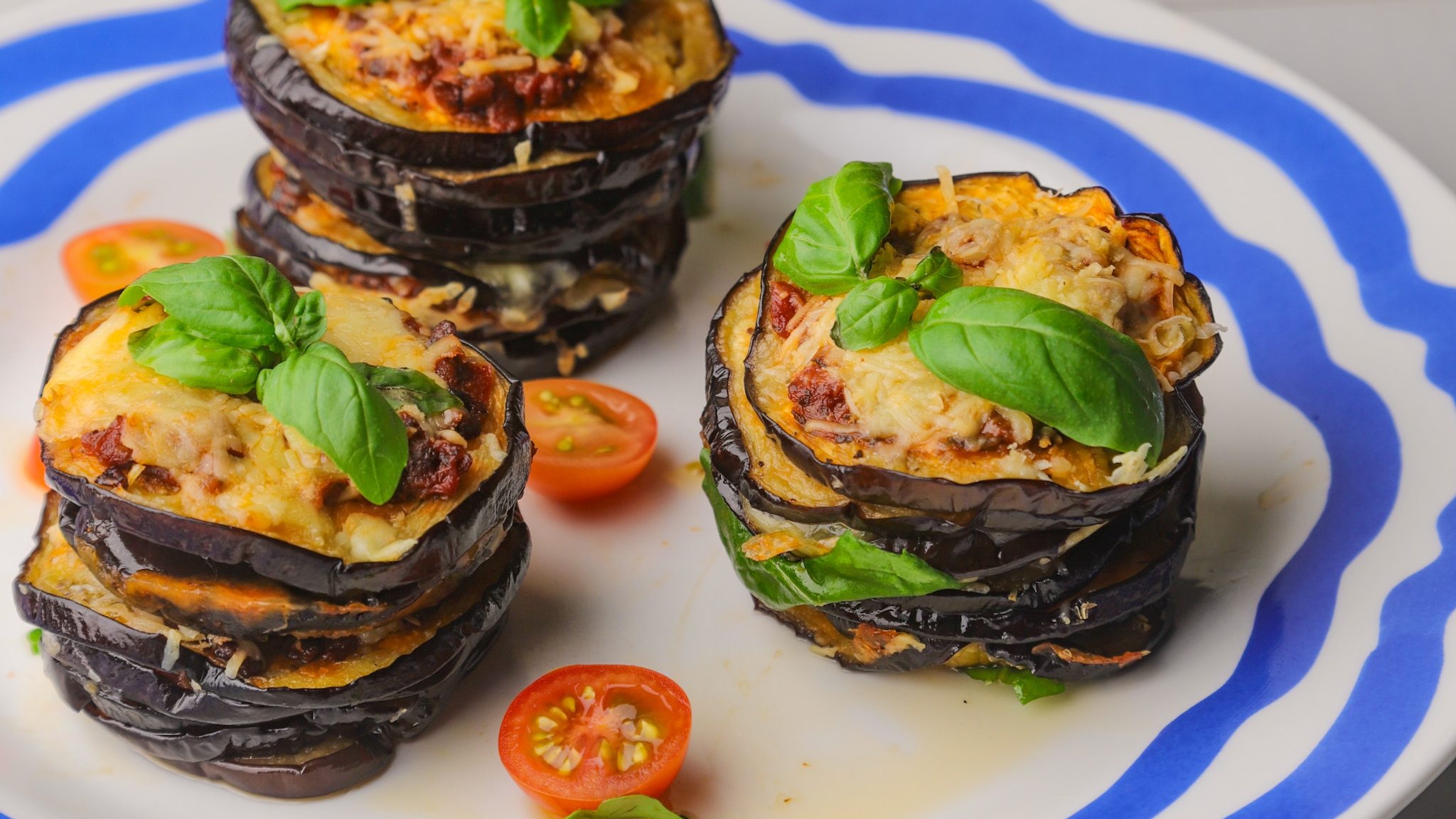 Three piles of aubergine slice stacks with basil leaves on top on a plate with cherry tomato halves.
