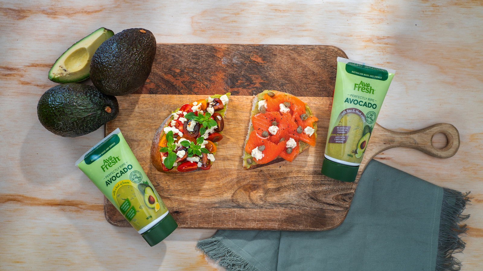 Two tubes of Avofresh avocado on a wooden board with two open topped sandwiches