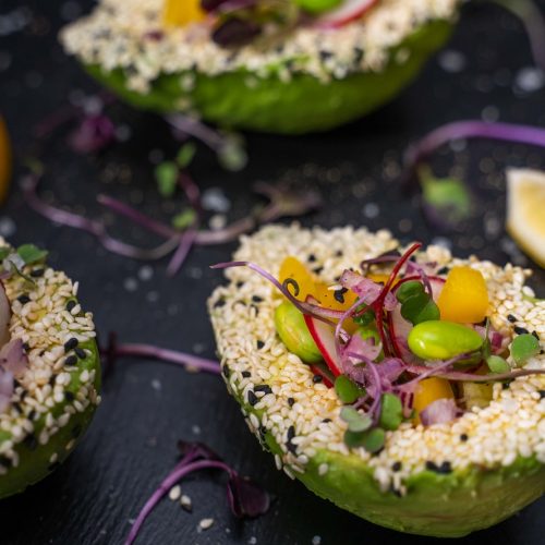 Two halves of avocado filled with colourful salad, topped with seeds on stone slate with lemon wedges.