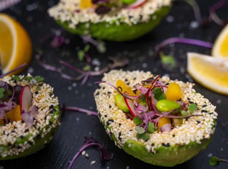 Two halves of avocado filled with colourful salad, topped with seeds on stone slate with lemon wedges.