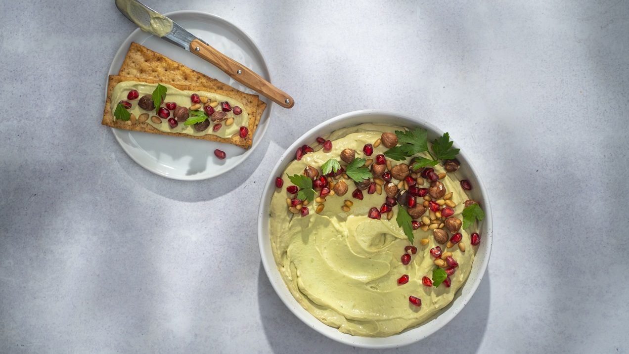 A bowl of pale green spread topped with colourful seeds, nuts and herb. A plate of crackers with dip spread over one and a knife.