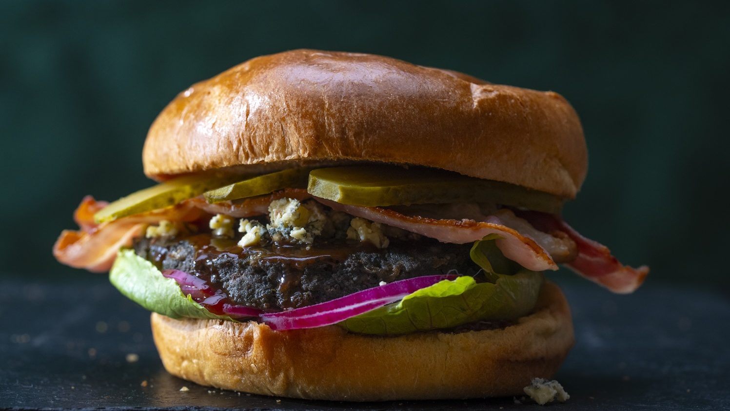 A beef burger with red onion, lettuce, pickles on black background.