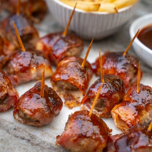 Several brown sauced bacon wrapped meatballs with picks on white board with potato fries and sauce.