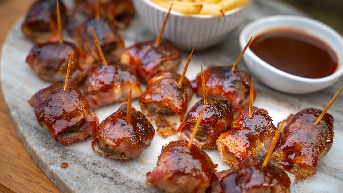 Several brown sauced bacon wrapped meatballs with picks on white board with potato fries and sauce.