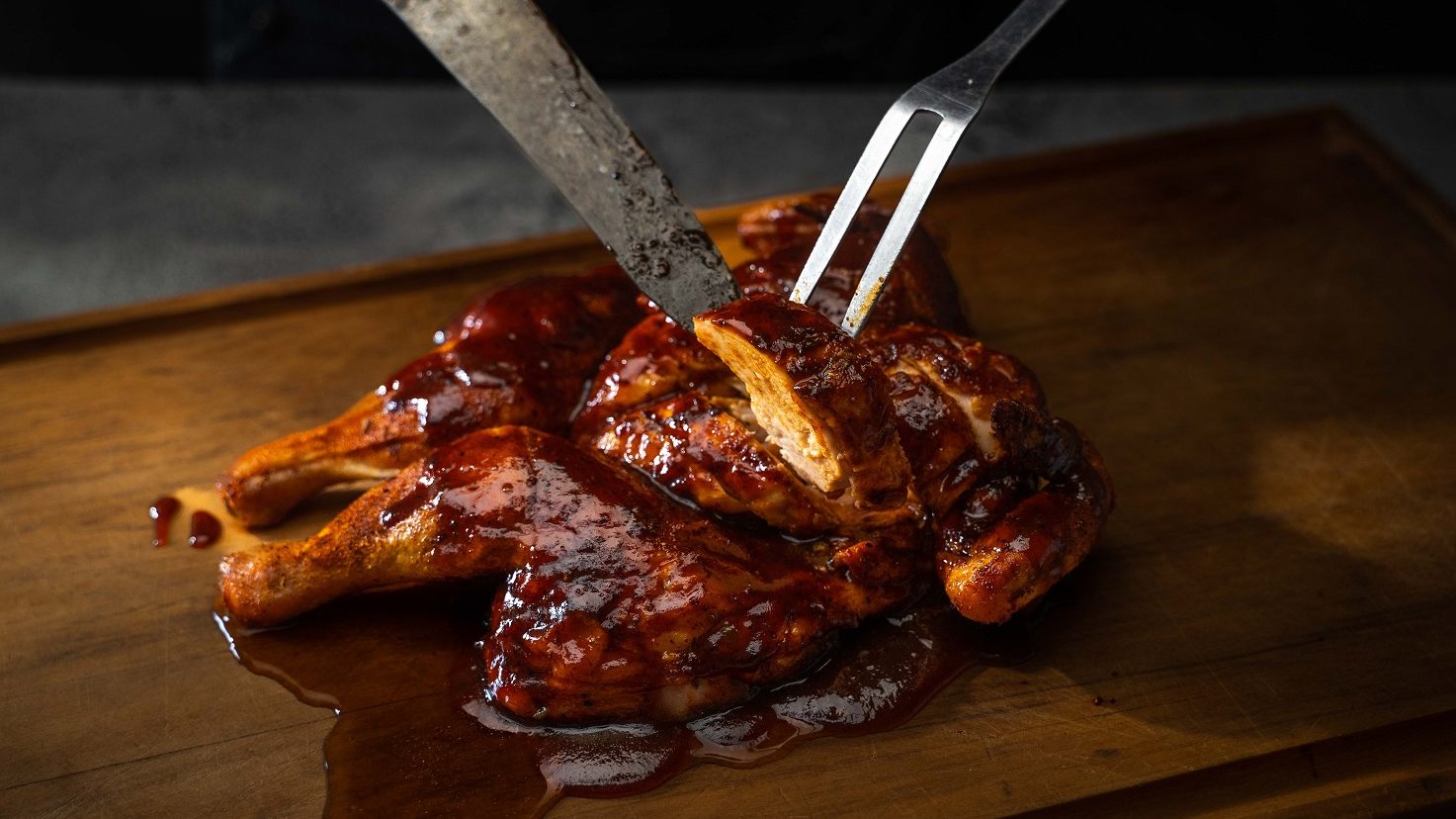 A cooked saucy whole chicken on a wooden board, carving knife and fork showing a sliced peice.