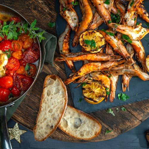 A plate of cooked large prawns and a pan of tomatoes with slices of bread and Christmas decorations.