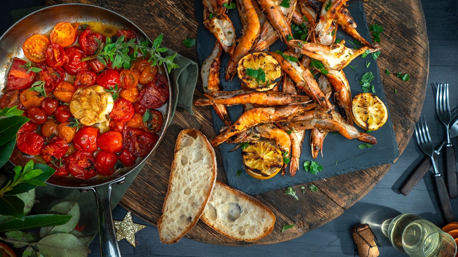 A plate of cooked large prawns and a pan of tomatoes with slices of bread and Christmas decorations.