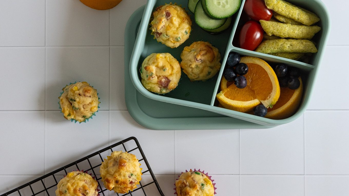 Three mini muffins in a green lunch box and four more muffins on wire rack.