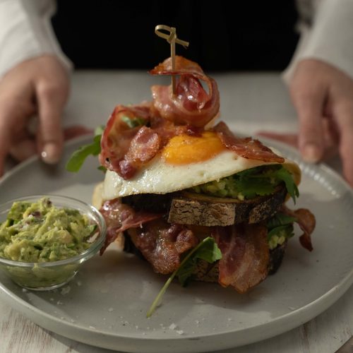 Bacon, bread, egg stack on a plate with a small bowl of green avo mash.