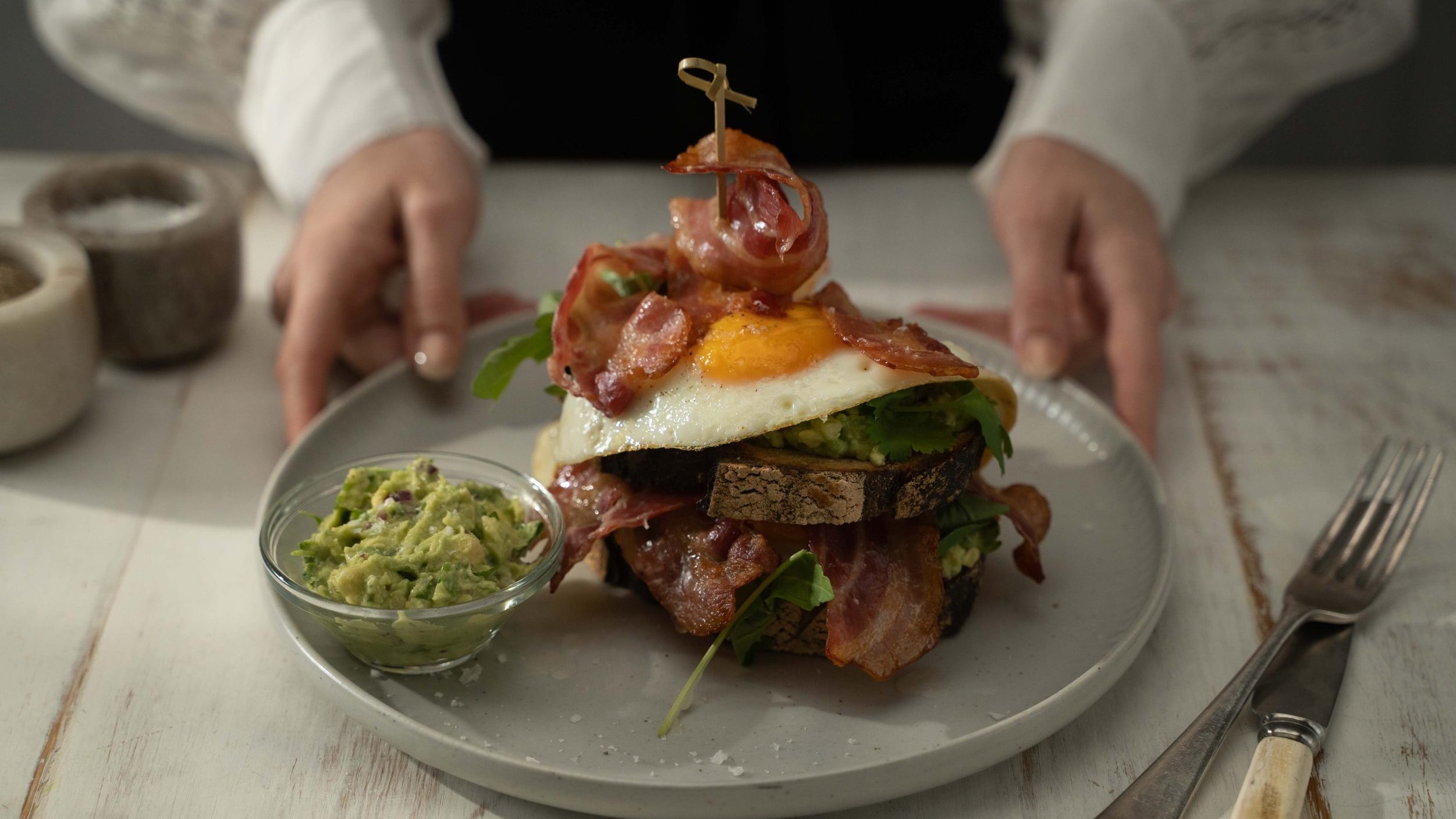 Bacon, bread, egg stack on a plate with a small bowl of green avo mash.