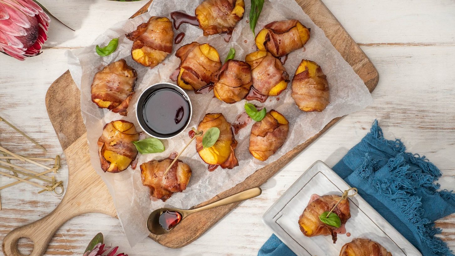Several bacon wrapped food and a pot of dark sauce, herb scattered on top on paper on wooden board , protea flowers