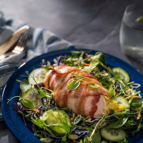 Bacon wrapped chicken breast on green salad on a blue plate, glass of water and knife and fork