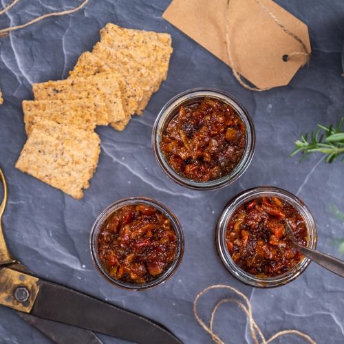 Trio of bacon jam jars on a platter, next to a fold of crackers and a vase of fresh rosemary.