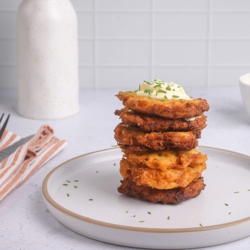 Six thin small brown fritters stacked on a white plate with a drop of white sauce on top on bench top.