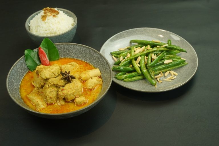 A bowl of coconut chicken curry, a plate of green beans and a small bowl of white rice on dark grey table.