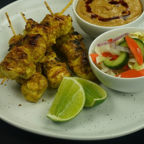Peanut satay - Four chicken skewers, bowls of salad and peanut sauce and lime wedges on a white plate of chicken