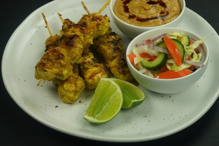 Peanut satay - Four chicken skewers, bowls of salad and peanut sauce and lime wedges on a white plate of chicken