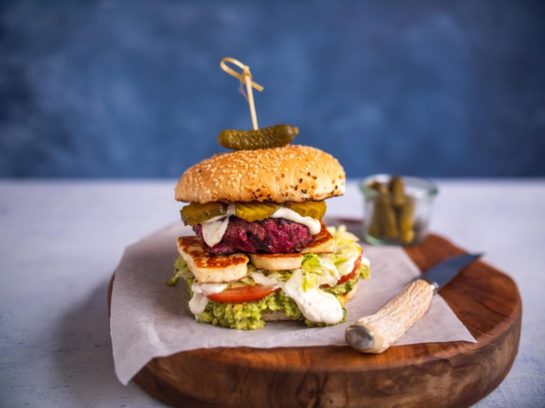 Beetroot burger generously garnished with avocado, mayo, halloumi cheese, tomatoes, lettuce and pickles placed a wooden cutting board.