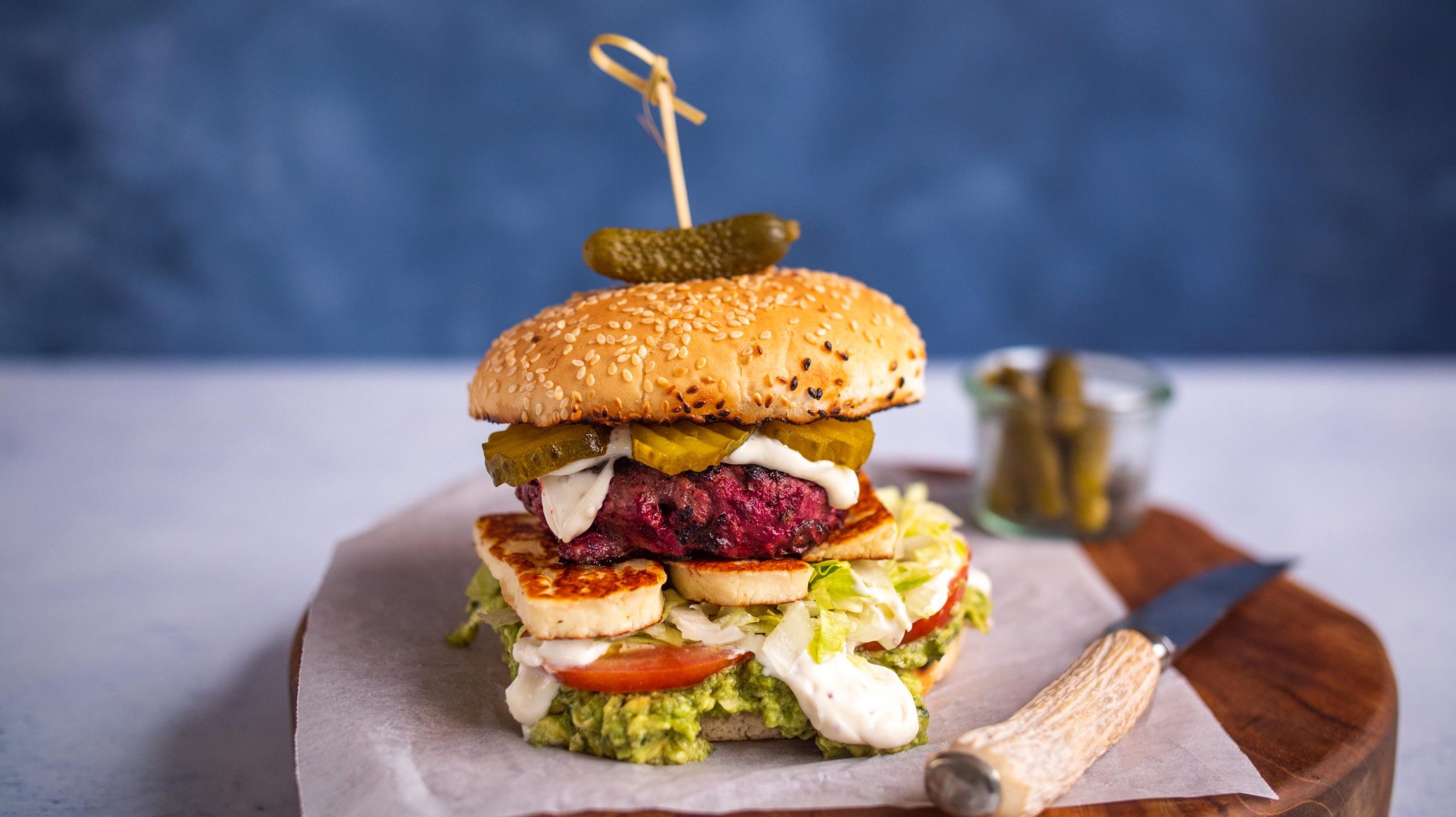 Beetroot burger generously garnished with avocado, mayo, halloumi cheese, tomatoes, lettuce and pickles placed a wooden cutting board.