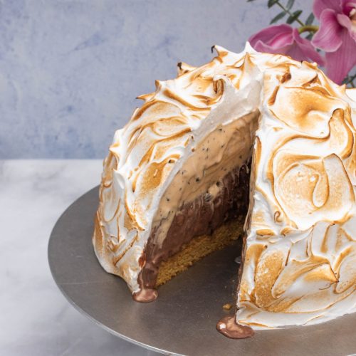 Gorgeous Bombe Alaska cake, also known as baked Alaska, with a slice taken out and ice cream filling slightly melting, set on a marble countertop with a bouquet of light pink flowers contrasting with the lightly torched meringue topping.