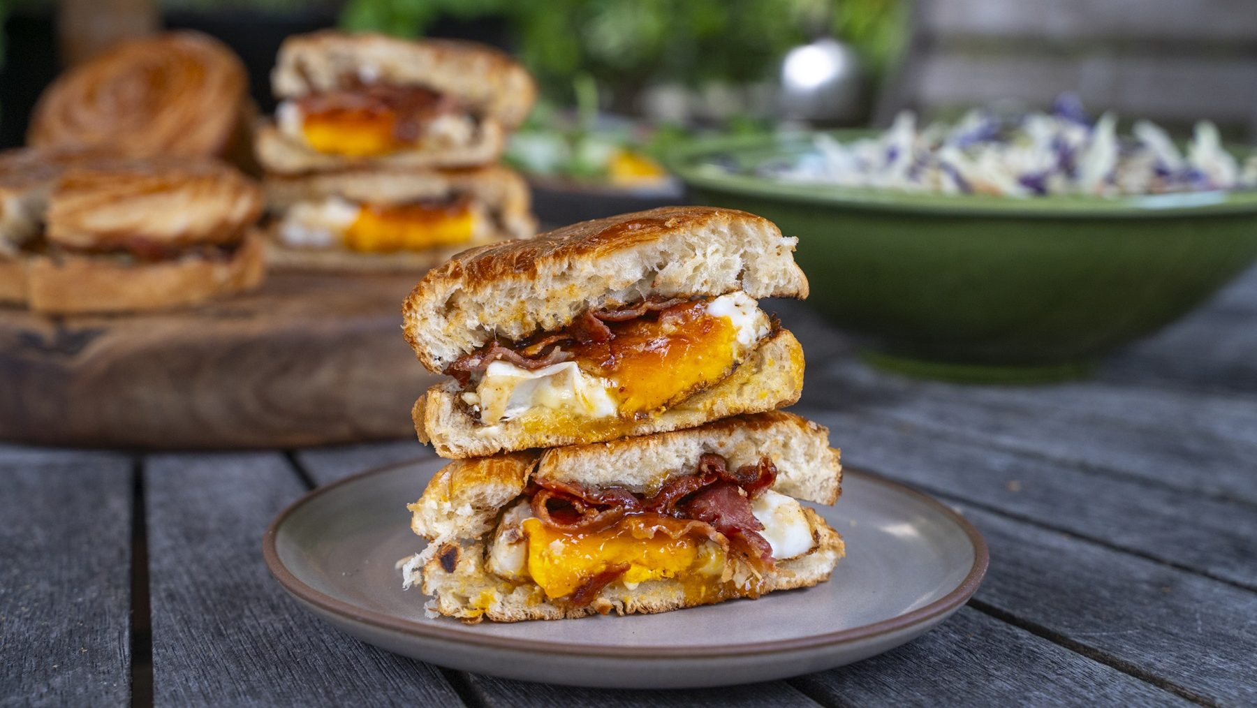 Two halves of a bun sandwich stack together on a plate, showing bacon and egg filling. More sandwich and a bowl of salad in background.