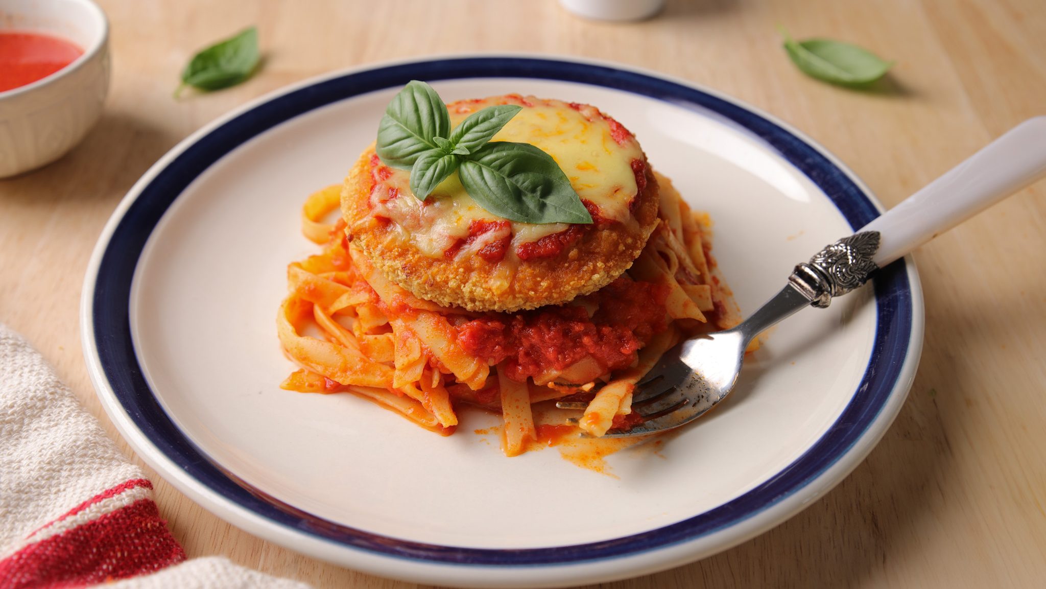 A piece of crumbed burger with melter cheese on top of tomato sauce pasta with basil on a plate and a fork.