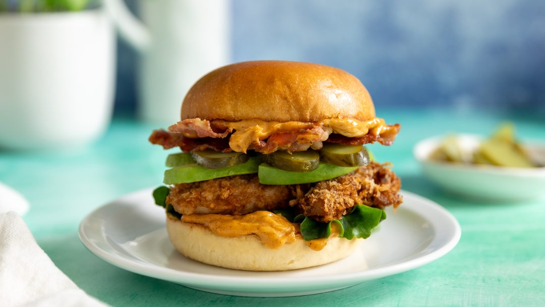 A side view of a fried chicken burger with avocado, bacon and gherkins.