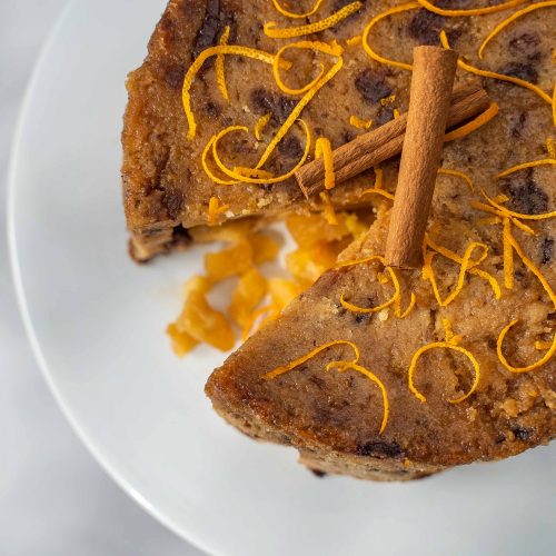 A large round cake with a wedge missing topped with cinnamon sticks and orange peels. apple stuffing showing in the gap on the white plate.