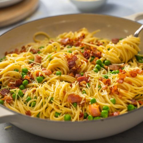 A white shallow casserole of sphagetti with bacon and peas.