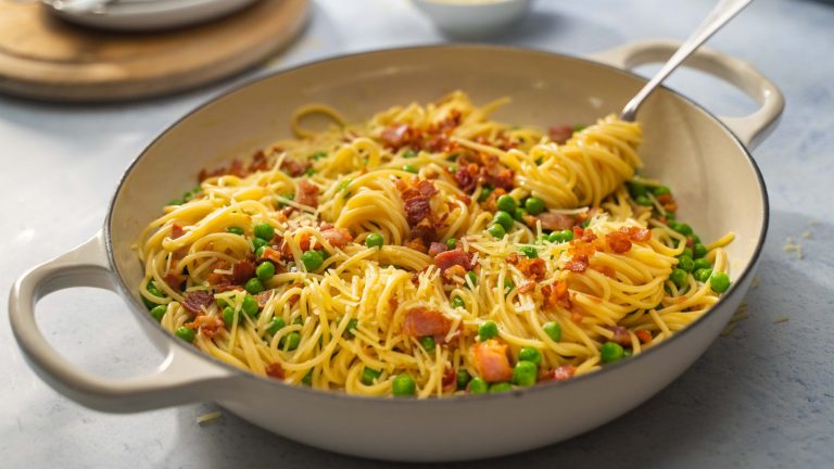 A white shallow casserole of sphagetti with bacon and peas.