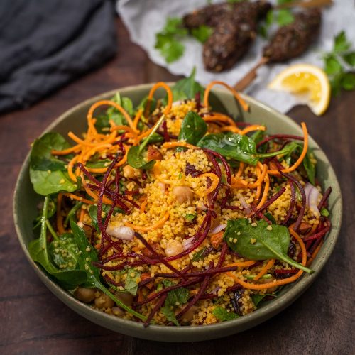 A bowl of yellow grains, red and orange strips and green leaf salad, meat kebabs and lemon wedges in the back.