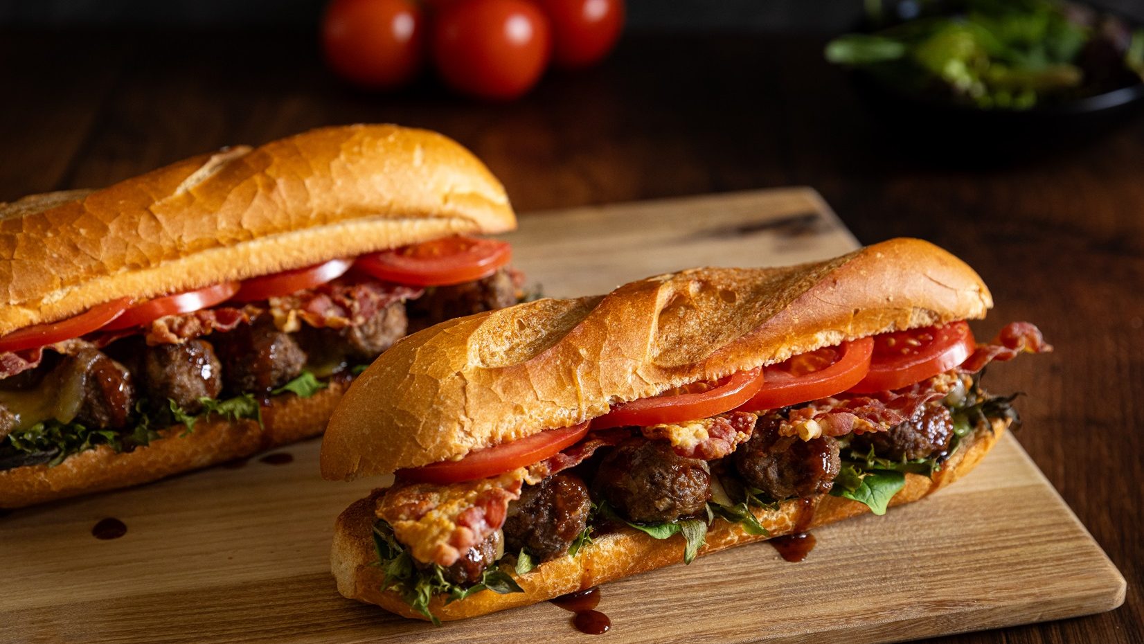 Two meatball baguette sandwiches on wooden board.