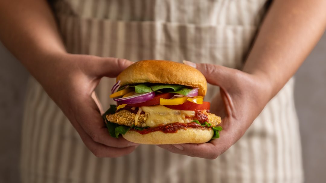 Two hands holding a burger filled with crumbed meat, tomato slice, red onion, cheese yellow capsicum and lettuce.