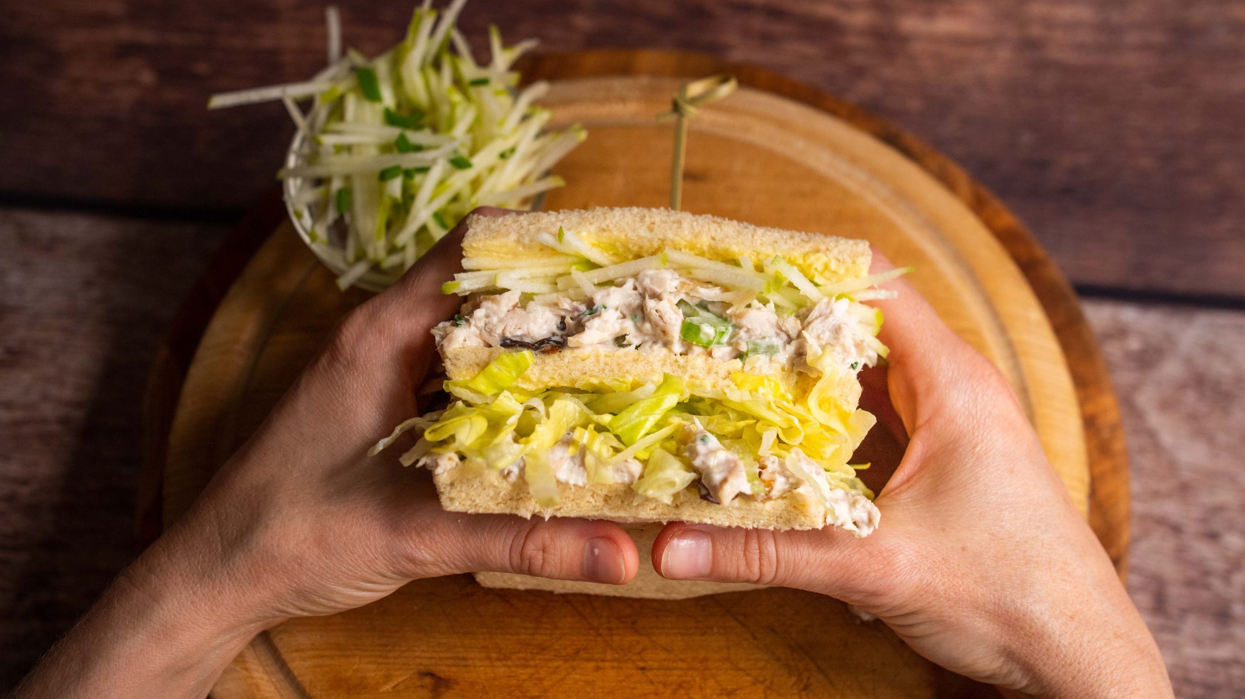 Chicken Waldorf salad club sandwich, close-up view held over a circular wooden cutting board, ready for a bite.
