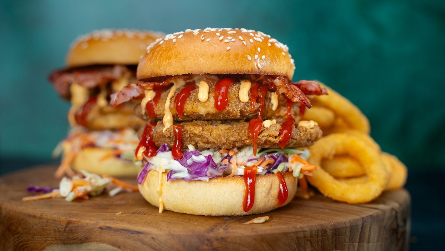 Two chicken burgers with slaw, bacon and drizzled sauces on a wooden board. Onion rings behind.