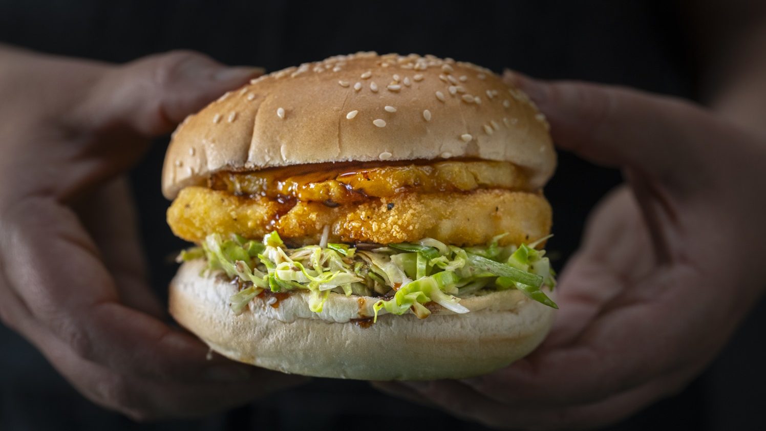 Hands holding a crumbed chicken burger with slaw and bacon .