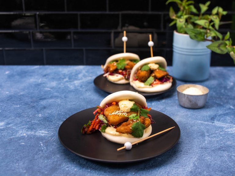 Three chicken karaage bao bun burgers with slaw and herb filling, served on black plates placed on a blue counter.