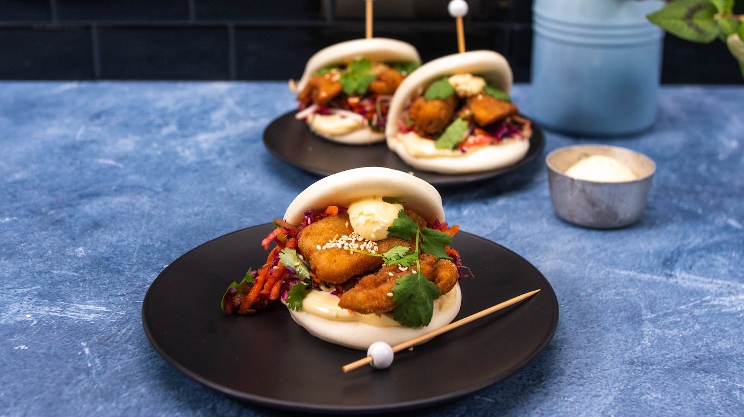 Three chicken karaage bao bun burgers with slaw and herb filling, served on black plates placed on a blue counter.